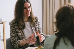 Pensive woman psychologist with brown hair in stylish clothes sitting and talking with unrecognizable female in light room about partial hospitalization services.