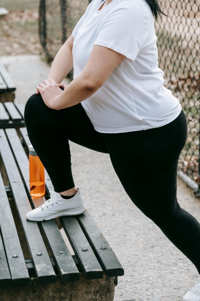 Crop anonymous plus size woman in white t shirt and leggings with sneakers exercising on bench while standing and stretching legs near water bottle in street near metal fence in daylight