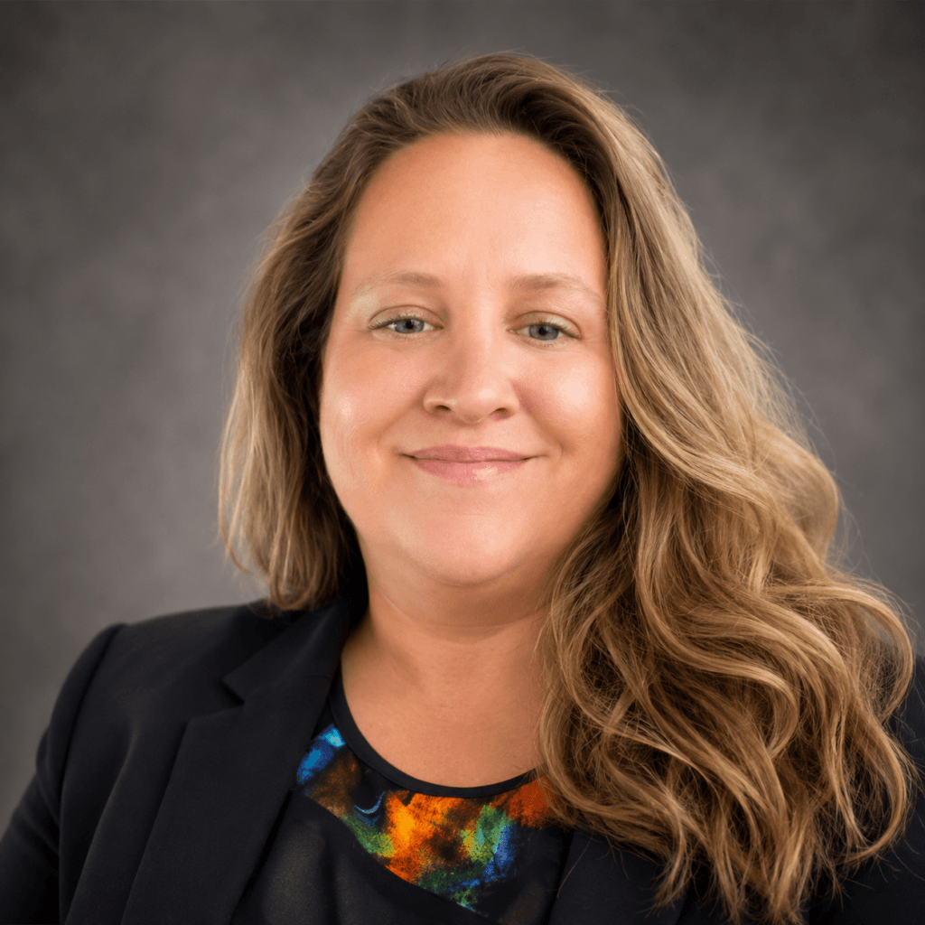 Alica Baxter A woman with long wavy blonde hair, wearing a black blazer and a colorful blouse, smiles softly at the camera against a neutral, textured background—her warmth reflects the compassion often found in substance abuse professionals.