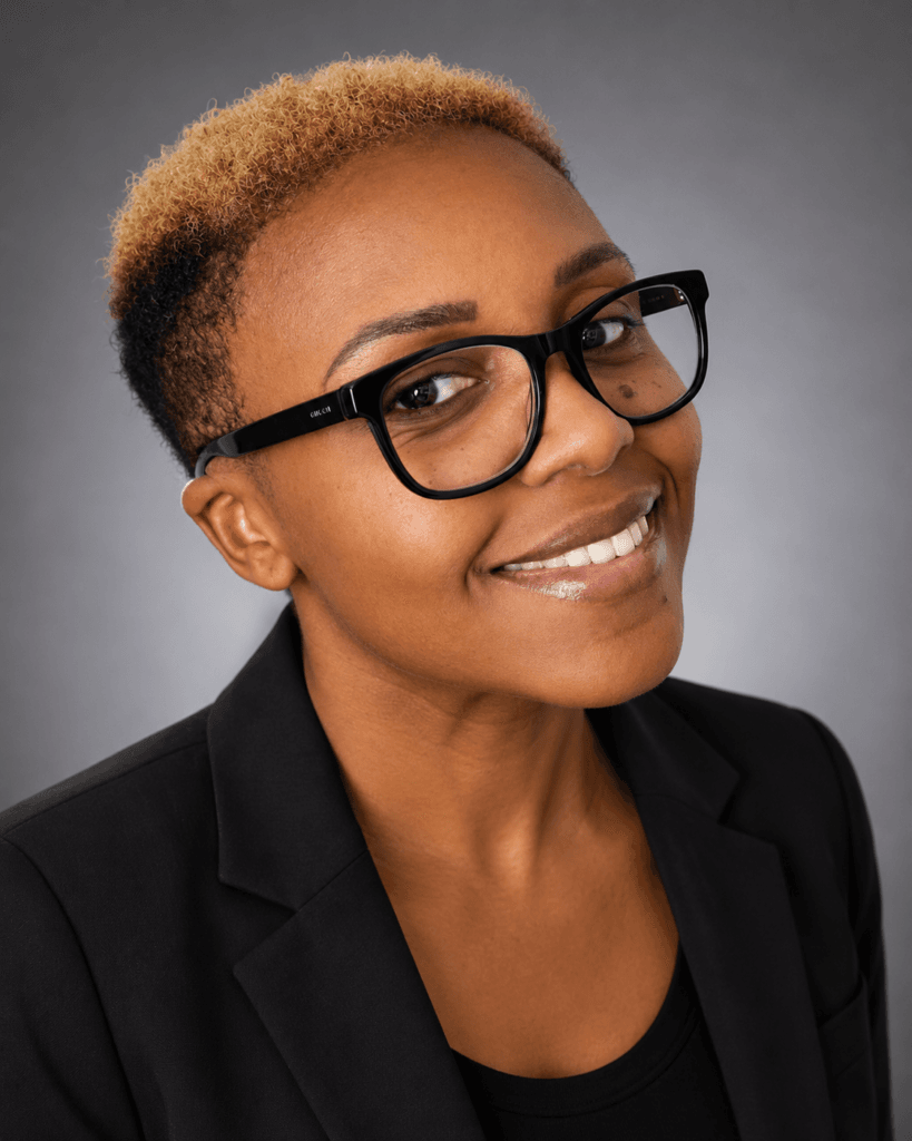 Lakeisha headshot Smiling addiction professional from Roanoke with short, curly, two-toned hair and black glasses, wearing a black blazer and top, poses in front of a neutral gray background.