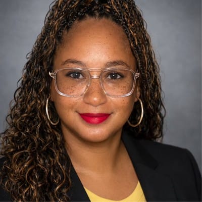 alicia bannister A woman with curly hair and hoop earrings, wearing clear glasses, a yellow top, and a black blazer, smiles in front of a gray background—embodying the approachable confidence of substance abuse professionals.