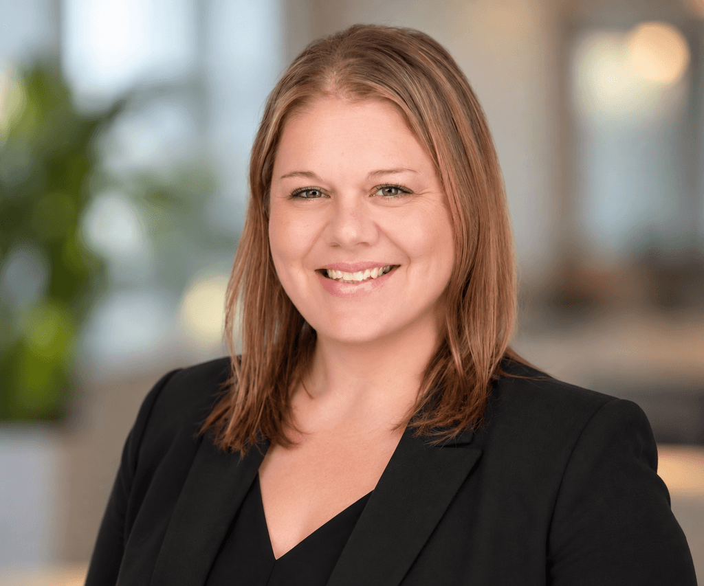 A woman with straight, light brown hair wearing a black blazer smiles at the camera. The background is softly blurred, with hints of greenery and indoor lighting, reflecting the calm professionalism often seen among substance abuse professionals.